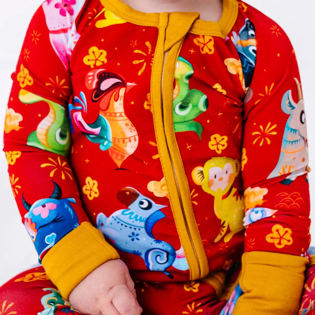 Close up of the front zipper and chest of a zip up baby pajama with mustard yellow trim and cuffs in a red print of zodiac animals and gold clouds.